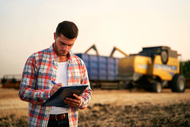 istock Farmer controls loading wheat from harvester to grain truck. Driver holding clipboard, keeping notes, cargo counting. Forwarder fills in consignment waybills. Agricultural commodities logistics 1262602166