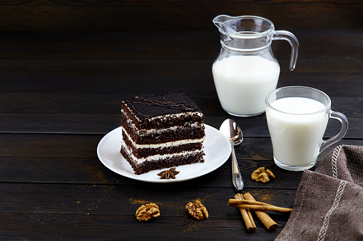 A piece of chocolate brownie cake with walnuts and a mug of milk on a dark wooden background.