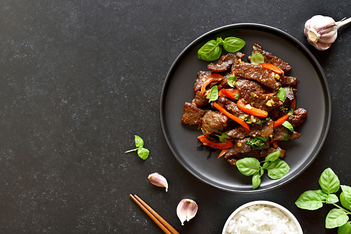 Thai beef stir-fry with pepper and basil on plate on a dark stone background with copy space. Top view, flat lay