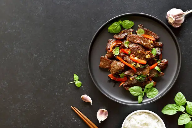 Thai beef stir-fry with pepper and basil on plate on a dark stone background with copy space. Top view, flat lay