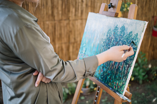 Female hand holding paintbrush and painting on canvas. Cropped woman painting picture on canvas on tripod in yard with blurred flowers in background.