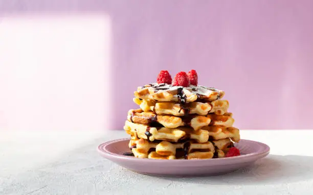 Stack of Viennese waffles with raspberries drizzled with chocolate topping in a pink plate. Summer breakfast on a lilac background with shadows.