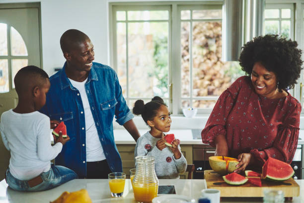 Smiling family eating a healthy snack of watermelon at home Smiling parents and their two adorable children standing at a kitchen counter at home and eating slices of watermelon black man eating watermelon stock pictures, royalty-free photos & images