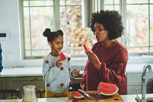 Mother and adorable little daughter sitting together at a kitchen counter at home and eating slices of watermelon