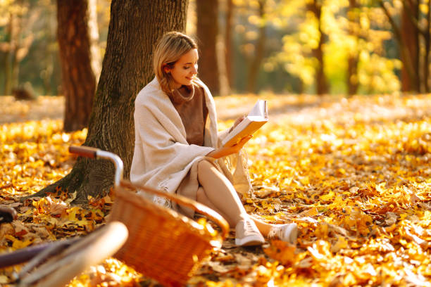 belle jeune femme s’asseyant sur un automne tombé laisse dans un parc, lisant un livre. - shawl photos et images de collection