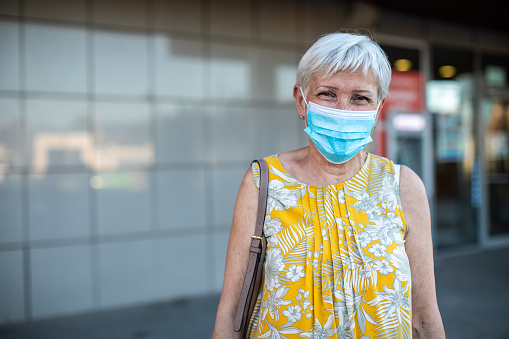 Portrait of senior woman with short, white hair, with protective face mask, standing in front of shopping mall, looking at camera and smiling