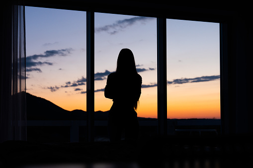 Silhouette of a young woman standing in front of a window with sunset in the background