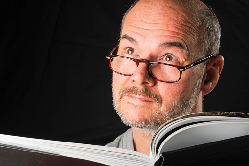 A mature man wearing glasses and a facial expression reads a book