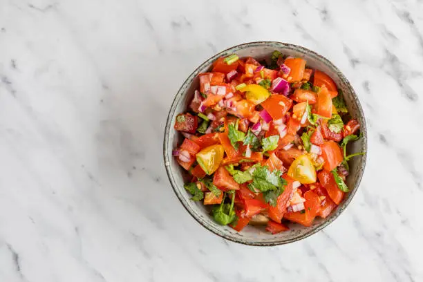 Tex Mex Pico de Gallo tomato salsa. The salsa contains tomatoes, cilantro and red onion. The bowl is seen from above with copy space, flat lay on a white marble background.