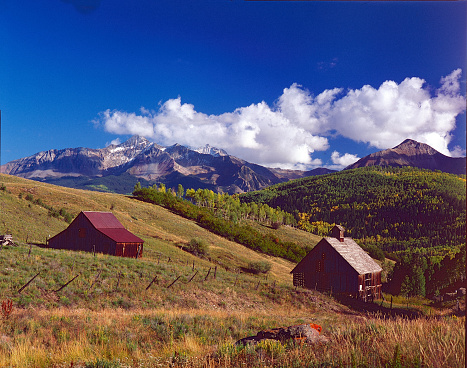 Scenic landscape in Colorado that shows an outbuilding and a barn with mountain peaks in the background and a beautiful blue sky.