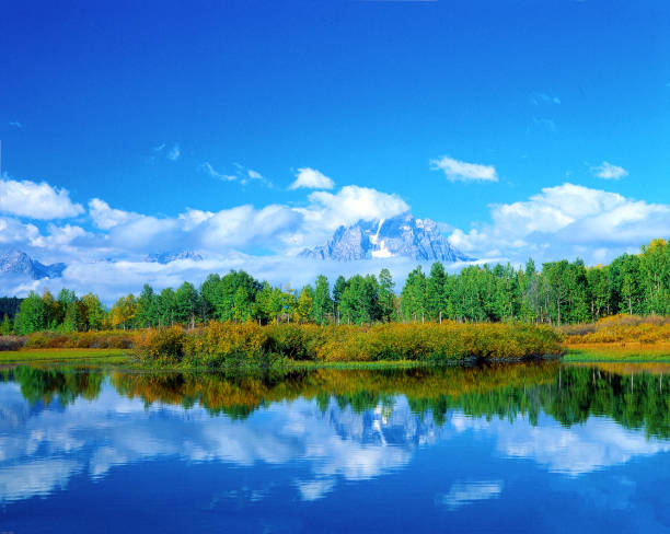 The calm water of Oxbow Lake in Wyoming reflects the shoreline of changing colors and the beautiful peaks of the Teton Mountains and the surrounding clouds.