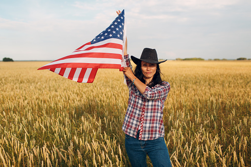 Woman farmer wearing cowboy hat, plaid shirt and jeans waving american flag at wheat field.