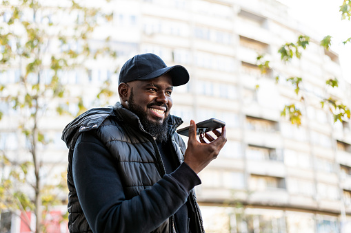 Mature African is walking in the city district and enjoying in his traveling adventure while talking over his mobile phone. A young bearded man of African-American ethnicity with a backpack and warm clothes is exploring the city.