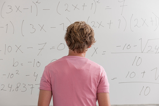 waist up rear view shot of a confused blond teenage boy  standing in front of whiteboard in classroom showing mathematical formula