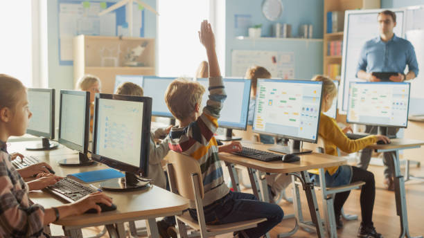 小学校の理科教室:教師は、パーソナルコンピュータで働くスマートな小学生を教育し、ソフトウェアコーディングのためのプログラミング言語を学びます。少年は質問で手を挙げる - elementary student little boys classroom child ストックフォトと画像