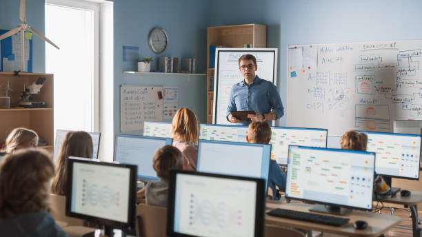 istock School Class: Portrait of the Enthusiastic and Motivated Teacher Using Digital Tablet Computer, Explains Lesson to Smart Children. Students getting Modern Education. 1262689361