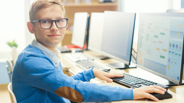 istock Elementary School Computer Science Classroom: Cute Little Boy wearing Glasses Uses Personal Computer, Learning Programming Language for Software Coding, Turned and Looks at Camera. 1262689411