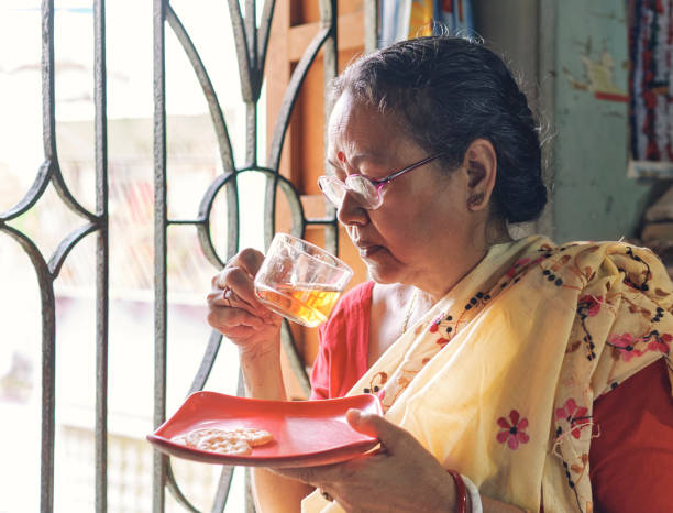 A simple looking middle aged Indian housewife drinking her morning cup of tea, sitting beside her room window. She is wearing a face mask. Concept of home quarantine.