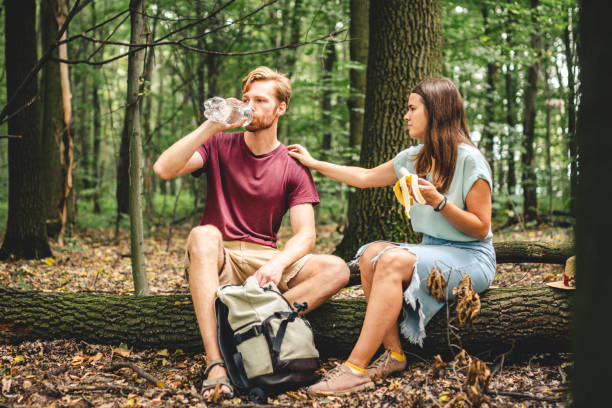 istock young couple sat to rest during forest walk along path to fallen tree, people drink water and eat banana fruit. Tourists break for snacks and replenishment water balance. Food and drinks in nature 1262691802
