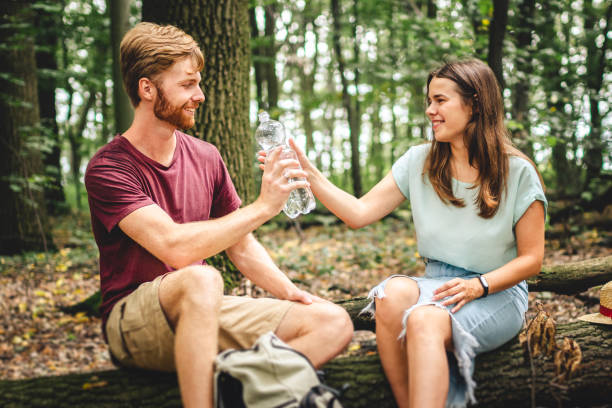 istock Caucasian couple stopped to rest while walking in the forest, young people sit on a fallen tree trunk drinking water from a bottle. Active rest, hike in a wooded area. Thirst and water balance 1262691981