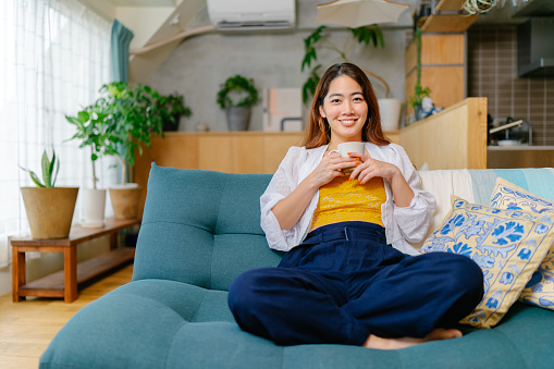 A young woman is sitting on a sofa in the living room holding a hot drink and smiling for the camera.