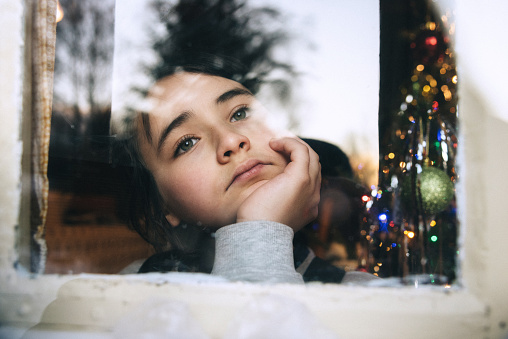 little girl looking out of window into the snowy winter landscape, in the background a shining decorated christmas tree
