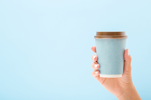 Young woman hand holding disposable paper cup on light blue background. Pastel color. Takeaway coffee or other drink. Closeup. Empty place for text. Front view.