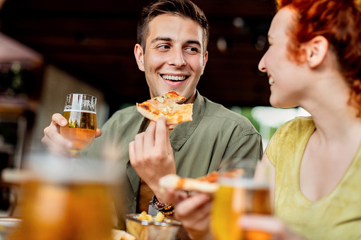 Young happy couple eating pizza and drinking beer while talking in a pub.