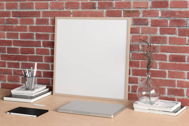 istock Square poster frame mockup on the wooden table of home studio workspace with red brick wall. Side view, clipping path around poster picture. 1262749899