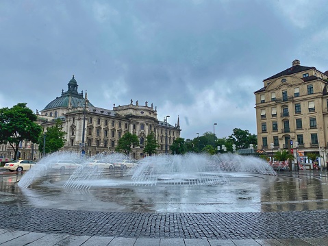 Old buildings, wet pavement and fountain at Karlsplatz (Stachus) square in Munich city center under the rain.