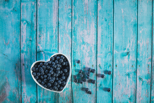Copy space shot of abundance of fresh blueberries in a heart shaped bowl, and some scattered around, lying on a blue wooden background.