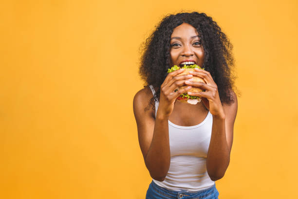 노란 배경에 고립 된 햄버거를 먹는 아프리카 계 미국인 흑인 아름다운 젊은 여성. - hamburger bun bread isolated 뉴스 사진 이미지