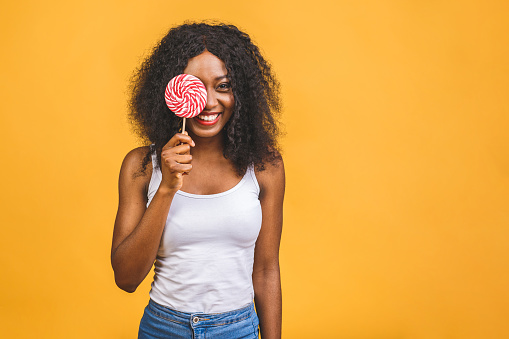 Smiling american african girl holding candy lollipop. Young cheerful woman with lollipop isolated over yellow background, copy space. Sweet life and confectionary.