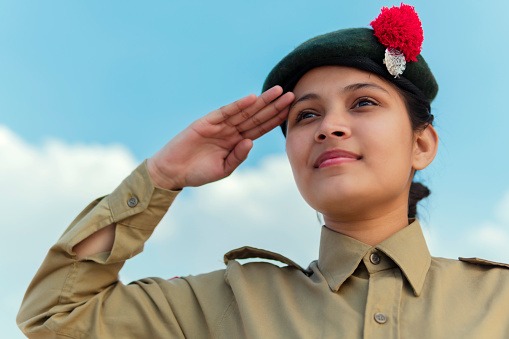 outdoor image of an happy Indian girl in NCC uniform, giving salute against blue cloudy sky and celebrating independence day (15 august).