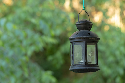 Old metal black painted candle lantern hanging in outdoor with trees in the background.
