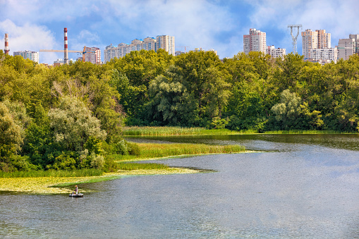 The picturesque landscape of the DniprÐ¾ Bay, a fisherman on a boat catches fish near the shore. The skyscrapers of the city's residential buildings are visible in the background.