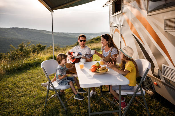 Happy family signing during their camping day by the trailer. Happy family having fun while singing at picnic table by the camper trailer in nature. Man is playing a guitar. motor home photos stock pictures, royalty-free photos & images
