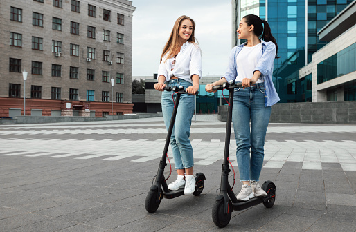 Full length portrait of two best female friends riding black motorized kick scooters in the city background