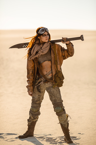 Post-apocalyptic woman warrior with weapon outdoors. Attractive fichter girl in shabby clothes holding sword standing in a confident pose looking away. People in nuclear post-apocalypse time. Life after doomsday concept. Desert and dead wasteland on the background.