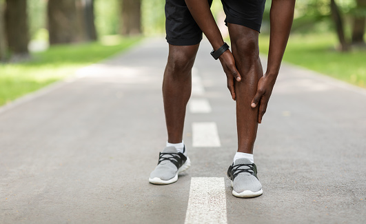 Closeup of black sportsman touching his aching leg while jogging outdoors, empty space