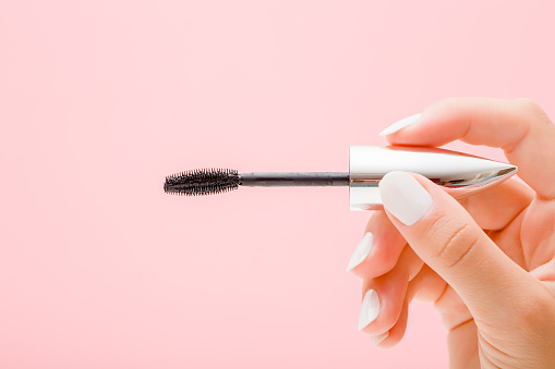 Young woman fingers holding black eyes mascara on light pink background. Pastel color. Female beauty product. Closeup.