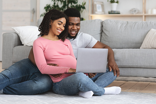 Loving pregnant african couple using laptop at home, sitting on floor at living room, watching baby room interior, empty space