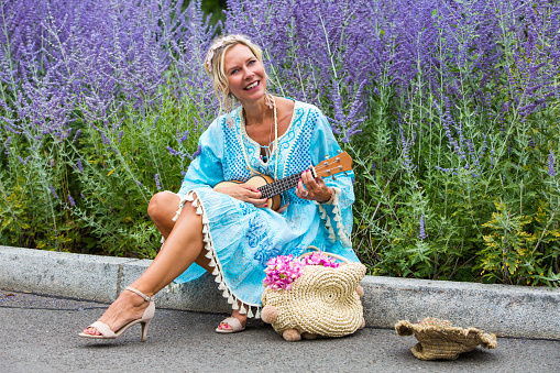 blond woman in her 40s sitting outdoor at curbside and playing guitar