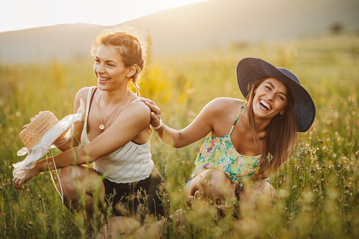 Two young cheerful female friends enjoying time together in nature, sitting in flower meadow and laughing