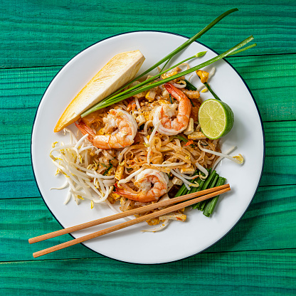 Prawn Pad Thai noodles with chopsticks. Prawn Pad Thai Noodles are a world-famous delicacy, here, this colorful traditional dish is photographed directly above on a round enameled metal white dish, with various condiments on the plate that are normally eaten together with the Pad Thai noodles. These condiments featured here are a raw banana flower, chives, bean shoots, and fresh lime. Sprinkling dried chili powder, sugar, and crushed peanuts on the dish is also very popular. This makes for a great color contrast composition with the dish set on an abstract, colorful, vibrant, worn, turquoise colored wood panel table background.