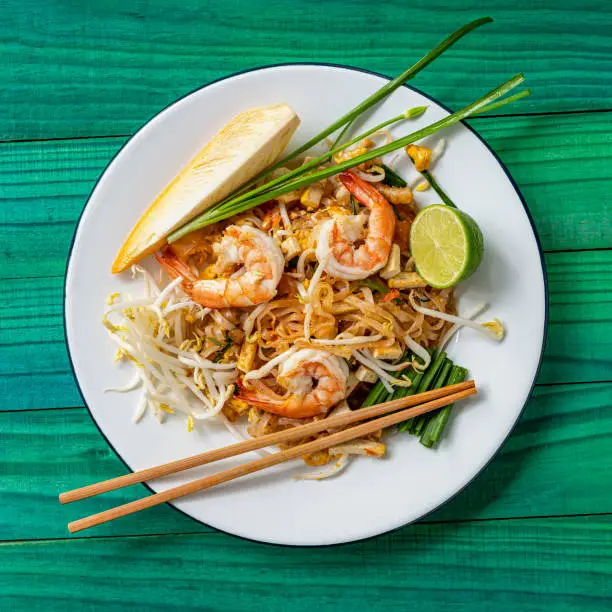 Photo of World famous freshly stir-fried cooked, Thai recipe of Prawn Pad Thai noodles on a round traditional enameled metal dish with chopsticks laid on the side of the dish, set on an abstract weathered turquoise colored wood panel table background.