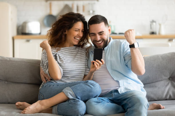 Overjoyed married couple celebrating online lottery win notification. Overjoyed married couple looking at smartphone screen, making yes gesture, celebrating online lottery win notification. Happy young spouses feeling excited about sms with good news, sitting on sofa. auction photos stock pictures, royalty-free photos & images