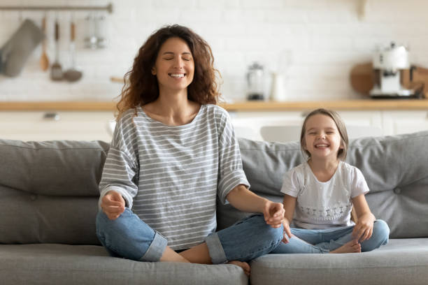 Happy young nanny mom teaching small daughter yoga breathing exercise. Full length happy young nanny mom sitting on sofa in lotus pose, teaching small daughter yoga breathing exercise. Friendly sincere two female generations family relaxing on couch, healthcare concept. mindfulness photos stock pictures, royalty-free photos & images