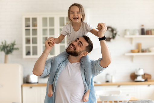 Cheerful dad carrying on neck playing with happy little preschool child daughter indoors. Playful small kid girl having fun with smiling bearded father at home, good family relations concept.