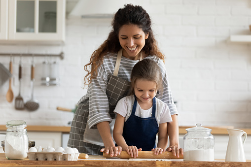 Happy attractive mommy helping cute smiling little preschool child daughter rolling dough for homemade pastry. Excited two female generations family enjoying cooking process together in kitchen.